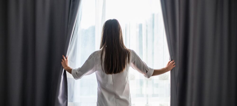 Eine Frau öffnet dunkle Vorhänge an einem Fenster. Darunter sieht man eine weiße, leichte Gardine.